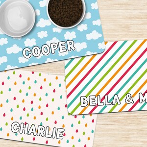 May include: Three personalized pet food mats with different designs. The first mat is blue with white clouds and the name "COOPER". The second mat is white with colorful raindrops and the name "CHARLIE". The third mat is white with colorful diagonal stripes and the names "BELLA & MAX".