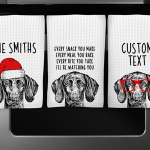 May include: Three white kitchen towels with black and white illustrations of a dog wearing a Santa hat, glasses, and a plain expression. The towels have text that reads "THE SMITHS", "EVERY SNACK YOU MAKE EVERY MEAL YOU BAKE EVERY BITE YOU TAKE I'LL BE WATCHING YOU", and "CUSTOM TEXT".