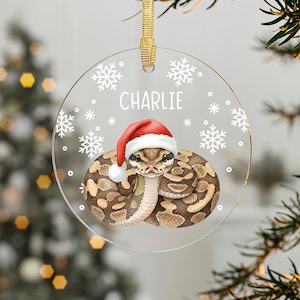 May include: A clear, round Christmas ornament with a gold ribbon. The ornament features a coiled snake wearing a Santa hat, with the name "Charlie" and snowflake designs. The snake has brown and tan markings.
