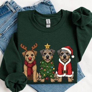 May include: Dark green sweatshirt featuring three dogs dressed for Christmas. One dog wears reindeer antlers and a scarf, another is a Christmas tree, and the third is in a Santa suit. The sweatshirt is displayed on a pair of blue jeans.