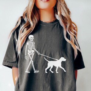 May include: A dark gray t-shirt with a white graphic of a skeleton walking a dog. The skeleton is wearing a leash and the dog is a simple outline.