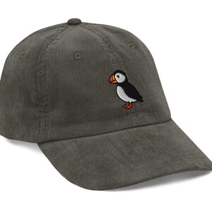 May include: A corduroy baseball cap in a muted green color. The cap features an embroidered puffin bird on the front. The puffin has a black body, white belly, orange beak and feet. The cap has a curved brim.