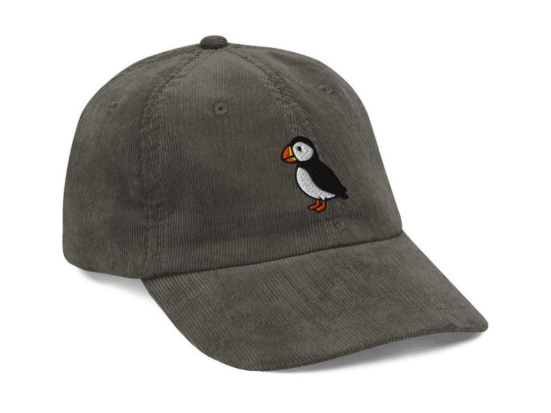 May include: A corduroy baseball cap in a muted green color. The cap features an embroidered puffin bird on the front. The puffin has a black body, white belly, orange beak and feet. The cap has a curved brim.