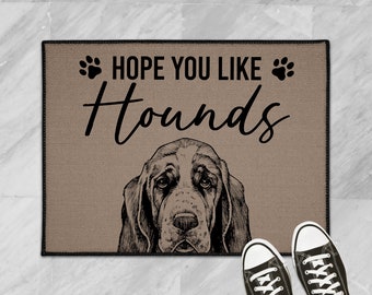 May include: A brown doormat with the text "HOPE YOU LIKE HOUNDS" in black lettering with paw prints on either side of the text. A black and white illustration of a Bloodhound dog's head is centered below the text.