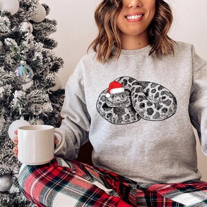Peut inclure: Un sweat-shirt gris clair avec une illustration détaillée d'un serpent portant un bonnet de Père Noël. Le serpent a un motif tacheté noir et blanc. Le sweat-shirt est associé à un pantalon de pyjama à carreaux rouges et noirs.