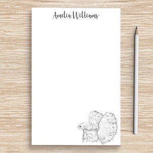 May include: A white notepad with the name "Amelia Williams" printed at the top. A black and white line drawing of a betta fish is at the bottom of the notepad.