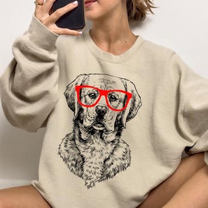 May include: A beige sweatshirt with a black and white illustration of a dog wearing red glasses. The dog is looking directly at the viewer.