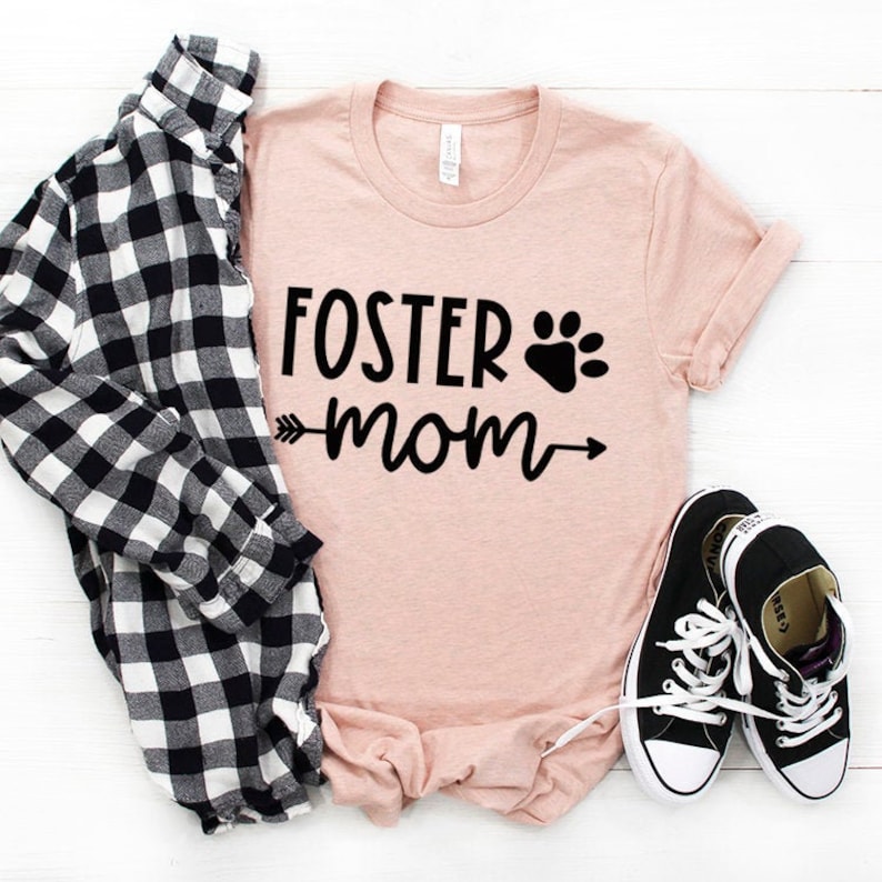 May include: A light pink t-shirt with the text "FOSTER mom" in black with a paw print and arrows on either side of the word "mom".