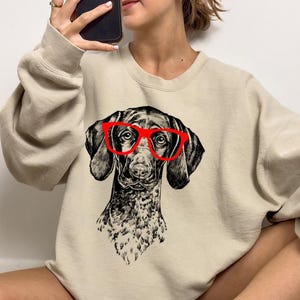 May include: A beige sweatshirt featuring a black and white illustration of a dog wearing red glasses.