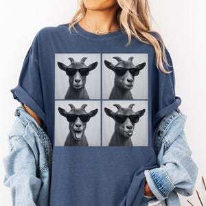 May include: A navy blue t-shirt featuring a square graphic of four goat portraits wearing sunglasses. The goats have different expressions. The shirt is worn with a light blue denim jacket.