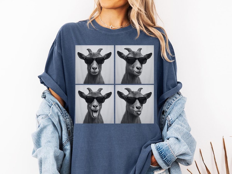 May include: A navy blue t-shirt featuring a square graphic of four goat portraits wearing sunglasses. The goats have different expressions. The shirt is worn with a light blue denim jacket.