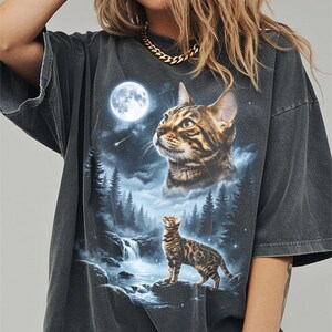 May include: Dark gray t-shirt featuring a detailed graphic of a cat gazing at a full moon above a forest and waterfall scene. The cat is brown and black with a spotted pattern. The shirt has short sleeves and a relaxed fit.