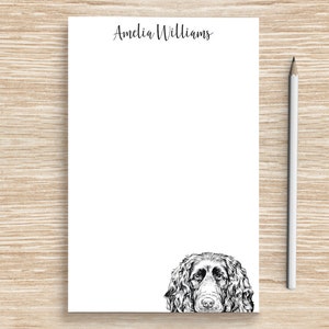 May include: White stationery with the name "Amelia Williams" in script at the top. A detailed black and white illustration of a dog's face is at the bottom. A white pencil rests on a light brown wooden surface.