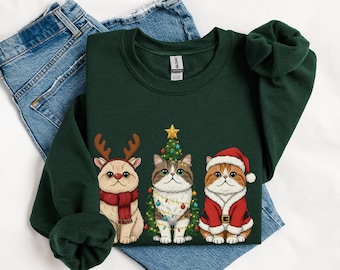 May include: A dark green sweatshirt featuring three cartoon cats dressed for Christmas. One cat wears reindeer antlers and a scarf, another is next to a Christmas tree, and the third is in a Santa suit. The sweatshirt is laid on top of blue jeans.
