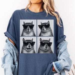 May include: A navy blue t-shirt featuring a four-panel image of a squirrel wearing black sunglasses. The squirrel's expressions vary in each panel, showcasing different moods. The shirt is a comfortable, casual style.