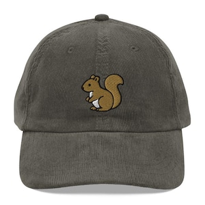May include: A corduroy baseball cap in a muted green color. The cap features an embroidered cartoon squirrel in shades of brown, tan, and white. The squirrel is centered on the front of the cap. The cap has a curved brim and an adjustable strap.