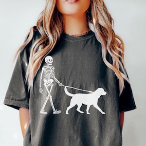 May include: A dark gray t-shirt with a white graphic of a skeleton walking a dog. The skeleton is wearing a leash and the dog is a labrador retriever.
