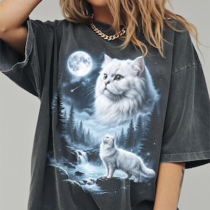 May include: Dark gray oversized t-shirt featuring a detailed graphic of a white cat under a full moon, with a waterfall and forest scene. The cat is looking up at the moon. The shirt has a vintage, washed look.