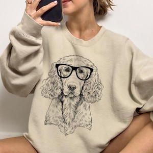 May include: A beige sweatshirt featuring a black and white illustration of a dog wearing glasses. The dog has long, curly hair and is looking directly at the viewer.