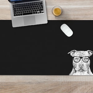 May include: Black desk mat with a white illustration of a dog wearing glasses. The dog is looking directly at the viewer. The mat is on a wooden surface.