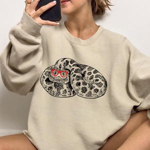 May include: A beige sweatshirt featuring a graphic print of a snake wearing red glasses. The snake is curled up and has a black and white pattern.
