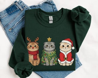 May include: A dark green sweatshirt featuring three cartoon cats dressed for Christmas. One cat wears reindeer antlers and a scarf, another is adorned with a Christmas tree, and the third is in a Santa suit.