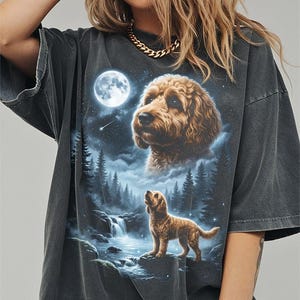 May include: Dark gray t-shirt featuring a detailed illustration of a golden doodle dog under a full moon. The design includes a waterfall, forest, and a second, larger dog head in the clouds. The shirt has a vintage, worn look.