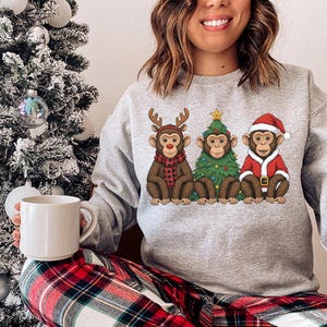 May include: A grey sweatshirt with a festive Christmas design. The graphic features three monkeys, one dressed as a reindeer, one as a Christmas tree, and one in a Santa Claus outfit.