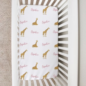 May include: White crib with a pink and white fitted sheet featuring a repeating pattern of giraffes and the name "Sophie".