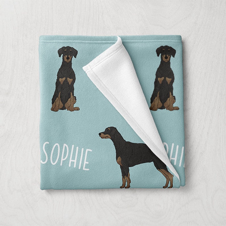 Puede incluir: Una manta de forro polar azul con un reverso de forro polar blanco. La manta presenta un patr&oacute;n repetido de perros Doberman Pinscher negros y marrones. El texto "Sophie" est&aacute; impreso en blanco en la manta.