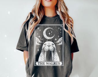 May include: A dark grey t-shirt featuring a white graphic of a walrus with a crescent moon and stars. The text "THE WALRUS" is printed below the image.