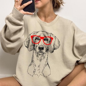 May include: A beige sweatshirt with a black and white illustration of a dog wearing red glasses.