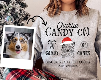 May include: A light gray sweatshirt with the text "Charlie Candy Co." and a dog wearing a Santa hat. The sweatshirt also features candy cane graphics and the words "Gingerbread & Hot Cocoa - Made Fresh Daily."