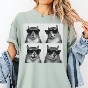 May include: A light green t-shirt featuring a four-panel image of a squirrel wearing sunglasses. The squirrel's expressions vary in each panel, with one showing a smile and another with its tongue out. The shirt is a comfortable, casual style.