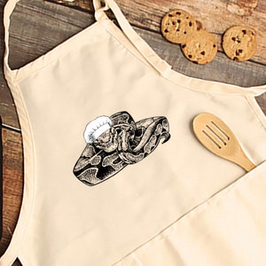 May include: Cream-colored apron with a black and white illustration of a snake wearing a chef's hat. The apron has a front pocket and a neck strap. Chocolate chip cookies and a wooden spoon are visible in the background.
