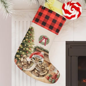 May include: Christmas stocking with a red and black buffalo plaid cuff. The stocking features a festive design with a Christmas tree, wreath, and a snake wearing a Santa hat with the name "Charlie". A red and white swirled lollipop is attached.