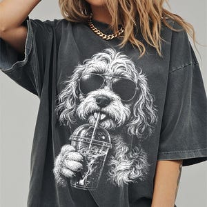 May include: A charcoal grey oversized t-shirt featuring a white graphic illustration of a curly-haired dog wearing dark sunglasses and drinking from a clear plastic cup with a straw. The shirt has a relaxed fit with short sleeves and a crew neckline.