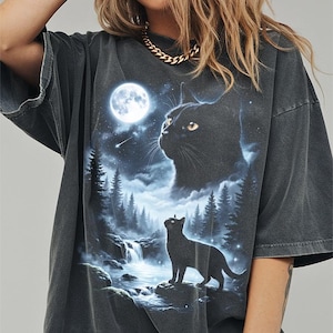 May include: Dark gray oversized t-shirt featuring a black cat design under a full moon. The graphic includes a waterfall, forest, and a second cat silhouette. The shirt has short sleeves and a relaxed fit.