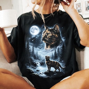 May include: Black t-shirt featuring a detailed graphic of a cat in a moonlit forest scene. The design includes a full moon, waterfalls, and pine trees. The shirt has a classic crew neck and short sleeves.