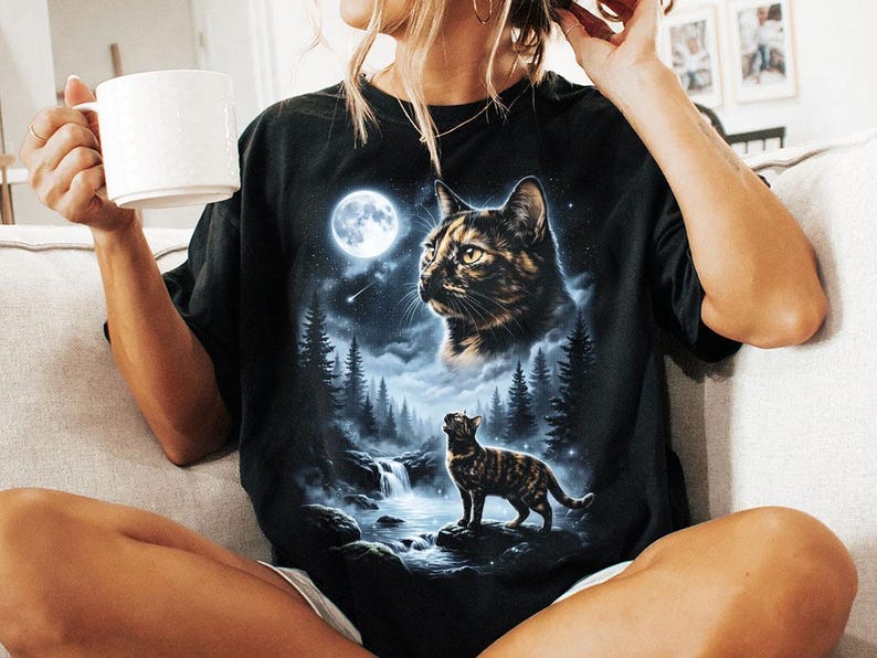May include: Black t-shirt featuring a detailed graphic of a cat in a moonlit forest scene. The design includes a full moon, waterfalls, and pine trees. The shirt has a classic crew neck and short sleeves.