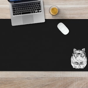 May include: Black desk mat with a white illustration of a hamster wearing glasses. The mat is on a light wood surface.