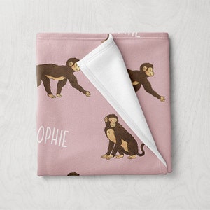 May include: A pink blanket featuring a repeating pattern of brown monkeys. The blanket has a white underside and the name "OPHIE" printed in white. The monkeys are in various poses.