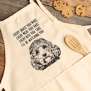 May include: Cream-colored apron with a pocket and a wooden spoon. The apron features a black and white illustration of a dog's face and the text: "EVERY SNACK YOU MAKE, EVERY MEAL YOU BAKE, EVERY BITE YOU TAKE, I'LL BE WATCHING YOU."
