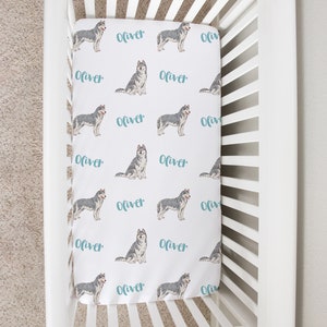 May include: White crib with a white fitted sheet featuring a repeating pattern of grey and white husky dogs and the name "Oliver" in teal blue.