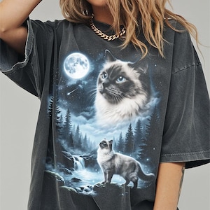 May include: Dark gray t-shirt featuring a detailed graphic of a cat in a moonlit forest scene. The design includes a full moon, a waterfall, and a second cat. The cat has blue eyes and a fluffy white chest.