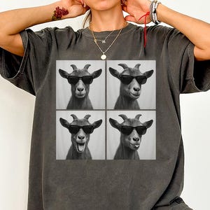 May include: Dark gray t-shirt featuring a four-panel image of a goat wearing sunglasses. The goat has different expressions in each panel. The shirt is a relaxed fit, with a crew neck and short sleeves.