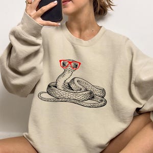 Peut inclure: Un sweat-shirt beige avec un motif de serpent portant des lunettes à monture rouge. Le serpent est enroulé et présente un motif détaillé.