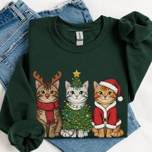 May include: Dark green sweatshirt featuring three cartoon cats dressed for Christmas. One cat wears reindeer antlers and a scarf, another is a Christmas tree, and the third is in a Santa suit. The sweatshirt is displayed on a pair of blue jeans.