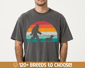 May include: A dark grey t-shirt with a retro-style sunset graphic featuring a Bigfoot walking a dachshund. The text "120+ Breeds to Choose!" is at the bottom of the image.