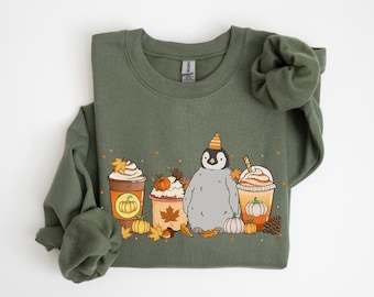 Op de afbeelding: Olijfgroene sweatshirt met een afbeelding van een pinguïn met een feesthoed, tussen drie herfst-thema koffiedranken. Het ontwerp bevat pompoenen, bladeren en een dennenappel, wat een herfstthema oproept.